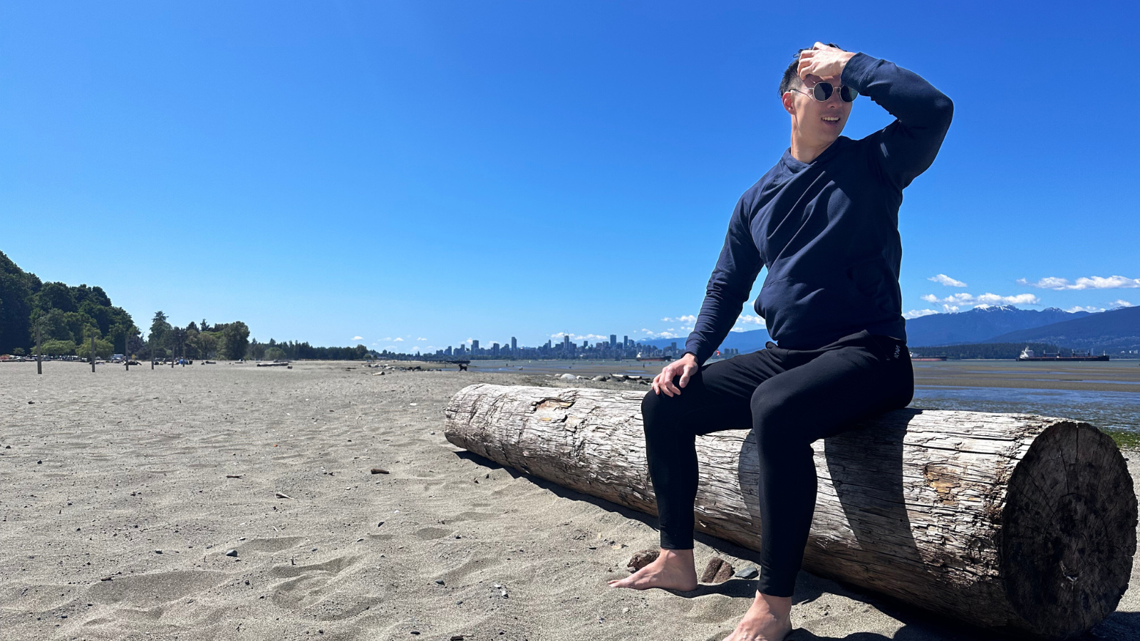 tall guy with sunglasses wearing navas lab navy sweatsuit. He is sat on a log at locarno beach