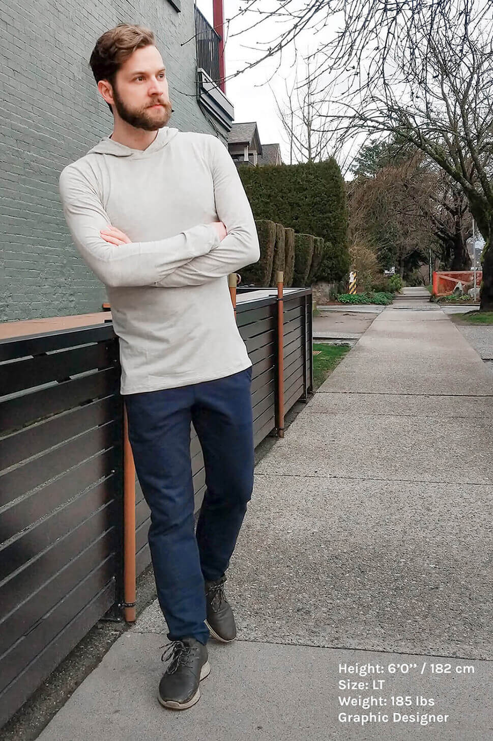 tall guy with beard wearing grey hooded t-shirt and navy pants leaning aginst fence