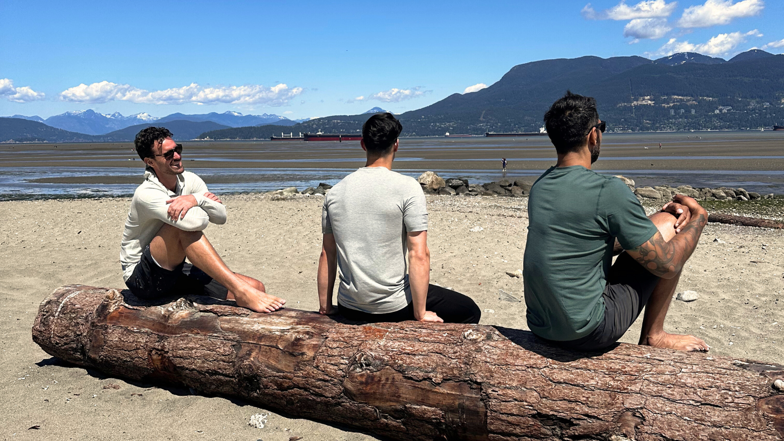 3 tall men sat on log at locarno beach looking at the ocean. They are all wearing navas lab casual t-shirts and shorts for tall men.