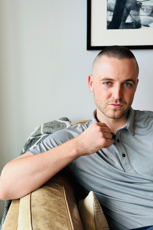 man with short hair wearing grey polo shirt by navas lab tall mens apparel. He is sat on leather couch