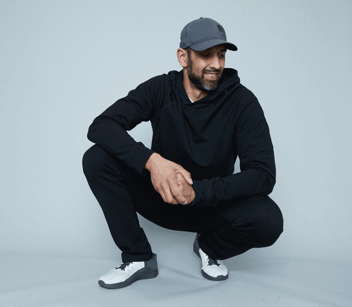 tall man wearing black navas lab hoody and sweatpants with grey hat, he is crouched down in grey studio